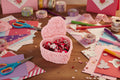 Pink heart-shaped candy box with rose patterns filled with red candies on a white background